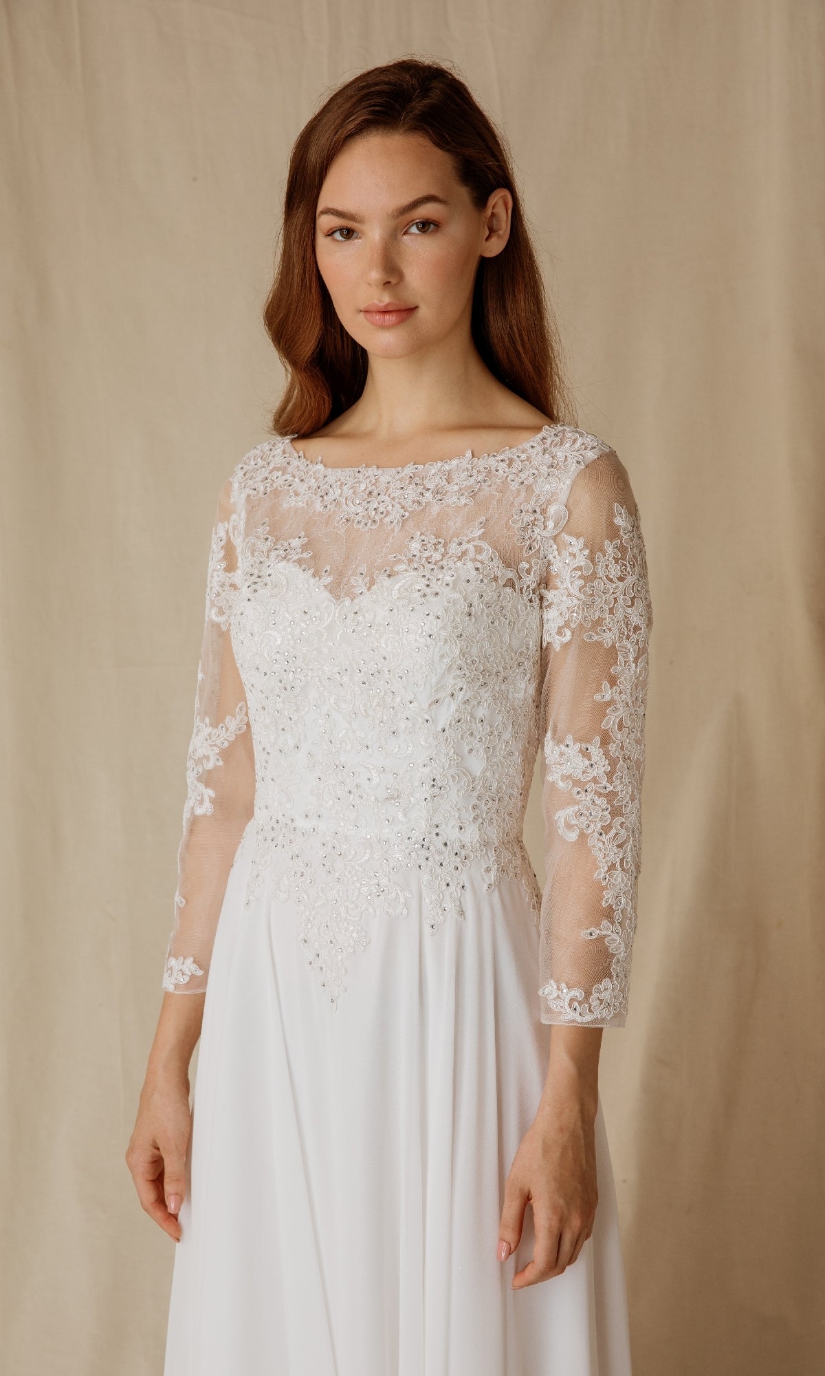 A woman stands before a beige backdrop in the Eureka 6909, a long formal dress featuring sheer long sleeves and delicate floral embroidery.